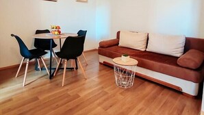 Una sala de estar con un sofá marrón, dos sillas negras, una mesa de centro redonda de madera y una mesa de comedor con sillas. Hay dos cuadros en la pared.