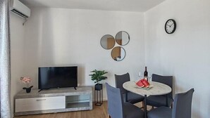 A modern living room with a flat-screen TV, a dining table with chairs, a small potted plant, and a wall clock.