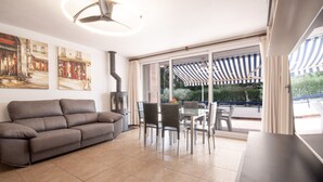 A living room with a sofa, dining table, chairs, and a large window with a view of a garden and a striped awning. - La Galera in Torredembarra (Torredembarra)