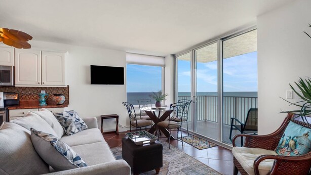 A modern living room with a sofa, coffee table, and a dining area with chairs. A flat-screen TV is mounted on the wall, and there is a large sliding glass door leading to a balcony with a view of the sky. - Estero Beach & Tennis 1206 A (Fort Myers Beach)