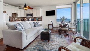 A modern living room with a sofa, coffee table, and dining area. A ceiling fan and kitchen are visible in the background. - Estero Beach & Tennis 1206 A (Fort Myers Beach)