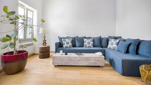 Ein Wohnzimmer mit einem blauen Ecksofa, einem Couchtisch, einer Topfpflanze und einem Fenster mit Tageslicht.