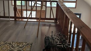 Ein geräumiges Dachzimmer mit einer Holzleiter, einem Schreibtisch mit elektronischen Geräten und großen Fenstern mit Blick auf Grünflächen.