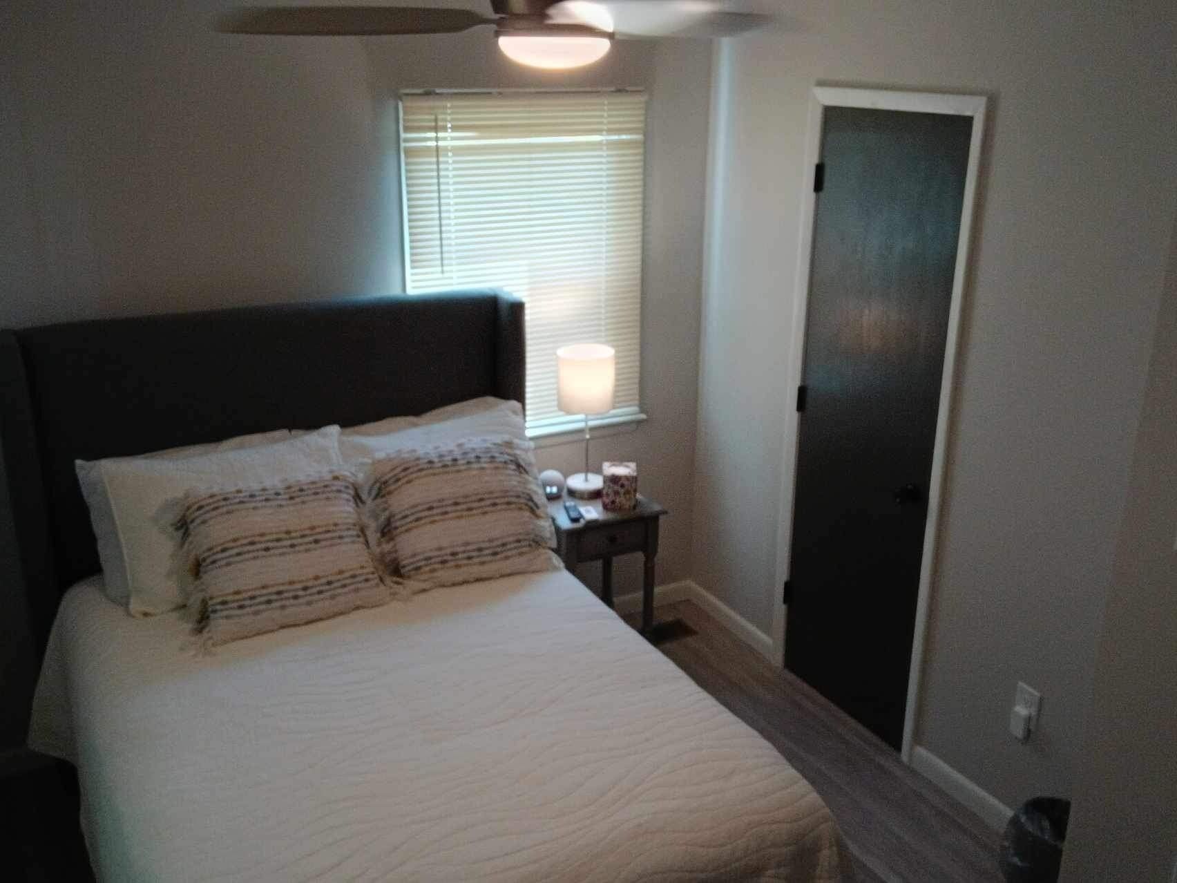 A bedroom with a bed, a nightstand, a lamp, a window with blinds, and a door.