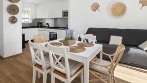 Une cuisine moderne avec des meubles blancs, une table à manger agrémentée de sets de table en osier et un canapé gris dans l’espace salon.