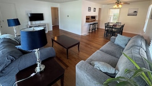 A living room with a sofa, coffee table, and lamp. There is a TV, a dining area with chairs and a table, and a kitchen area with a countertop and stools. - The Salon- 3 miles from WEC (Wilmington)