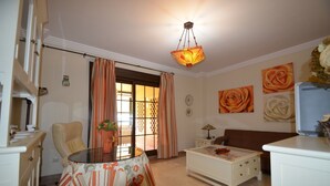 Een woonkamer met een bank, een salontafel, een stoel en een tafel met een bloemrijk tafelkleed. Gordijnen, een lamp en een plafondlamp zijn aanwezig.