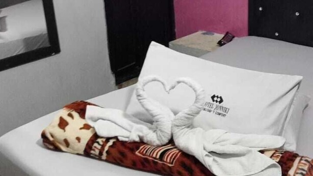 A neatly made bed with a heart-shaped pillow and a blanket, a bedside table with a book, a black headboard, and a wall-mounted fan. - Hotel Jhonyky (Bucaramanga)