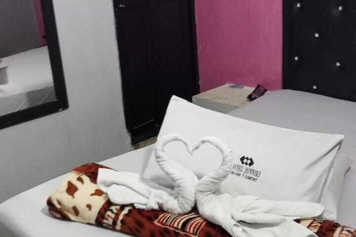 Uma cama bem arrumada com um travesseiro em formato de coração e um cobertor, uma mesa de cabeceira com um livro, um painel de cabeceira preto e um ventilador fixado na parede.