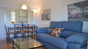 Una sala de estar con un sofá azul, una mesa de centro de vidrio, un rincón de comedor con sillas y una mesa, y un cuadro en la pared.