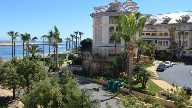 Un hotel de estilo arquitectónico mediterráneo, con una cúpula central y varios balcones. Se encuentra cerca de una playa con palmeras y un cielo azul despejado.