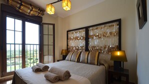 A hotel room with a large bed, two bedside lamps, a view of greenery through the window, and a decorative wall panel. - Los Pelicanos 94 - Two Bedroom Apartment, Sleeps 5 (Punta del moral)
