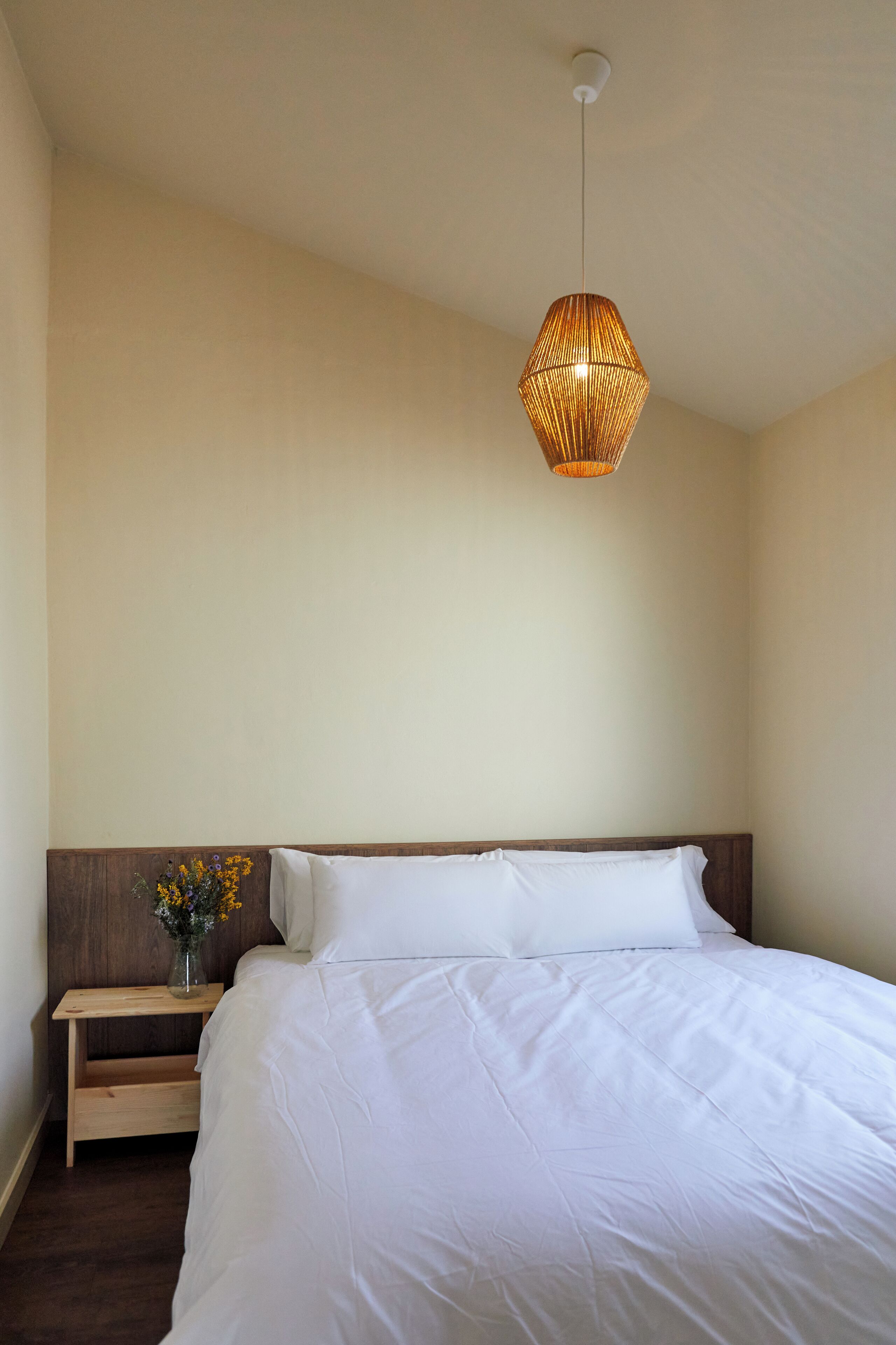 A bedroom with a large bed, a bedside table with a vase of flowers, and a hanging light fixture.