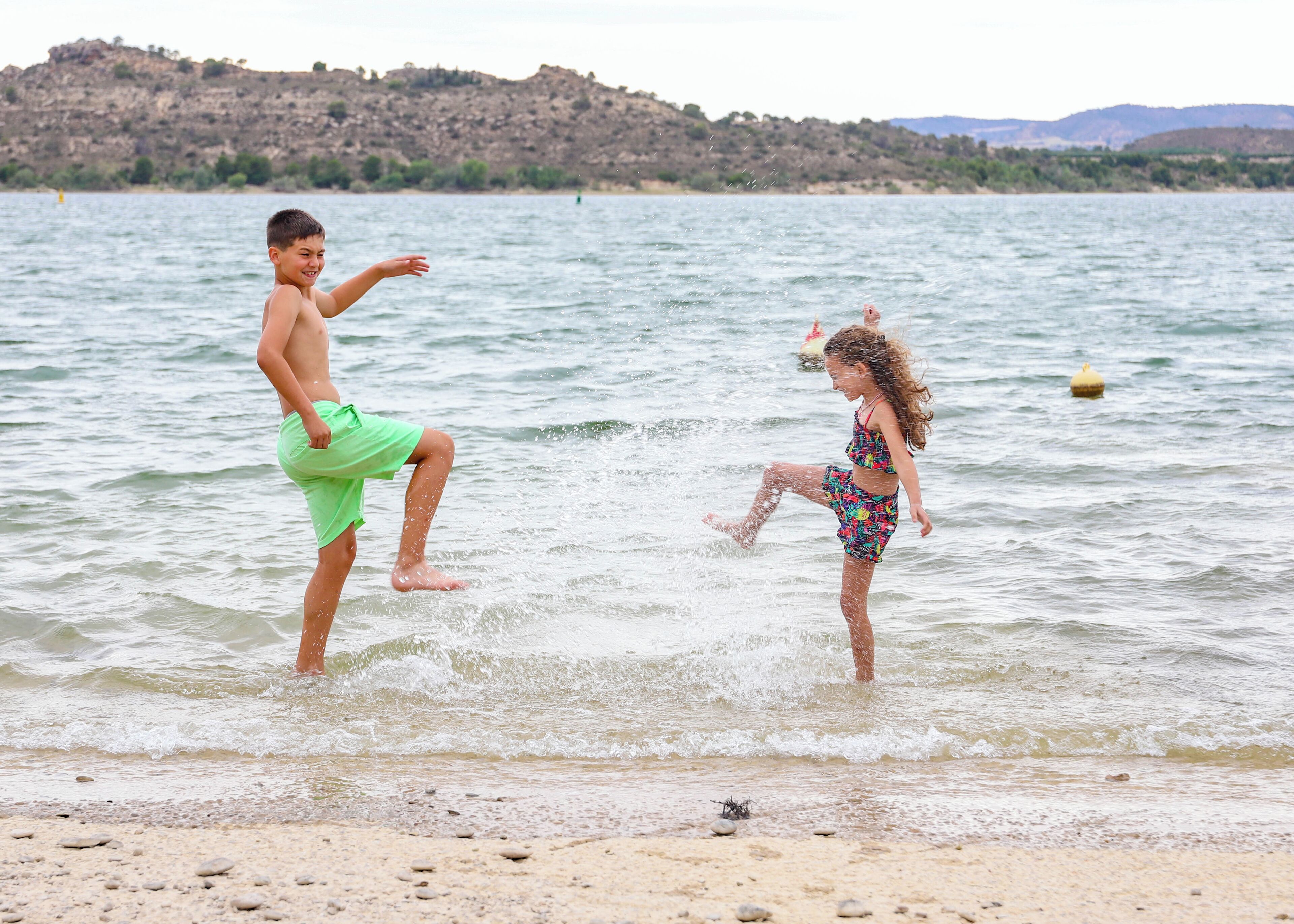 Two children playing in shallow water at a beach with a rocky landscape in the background.
