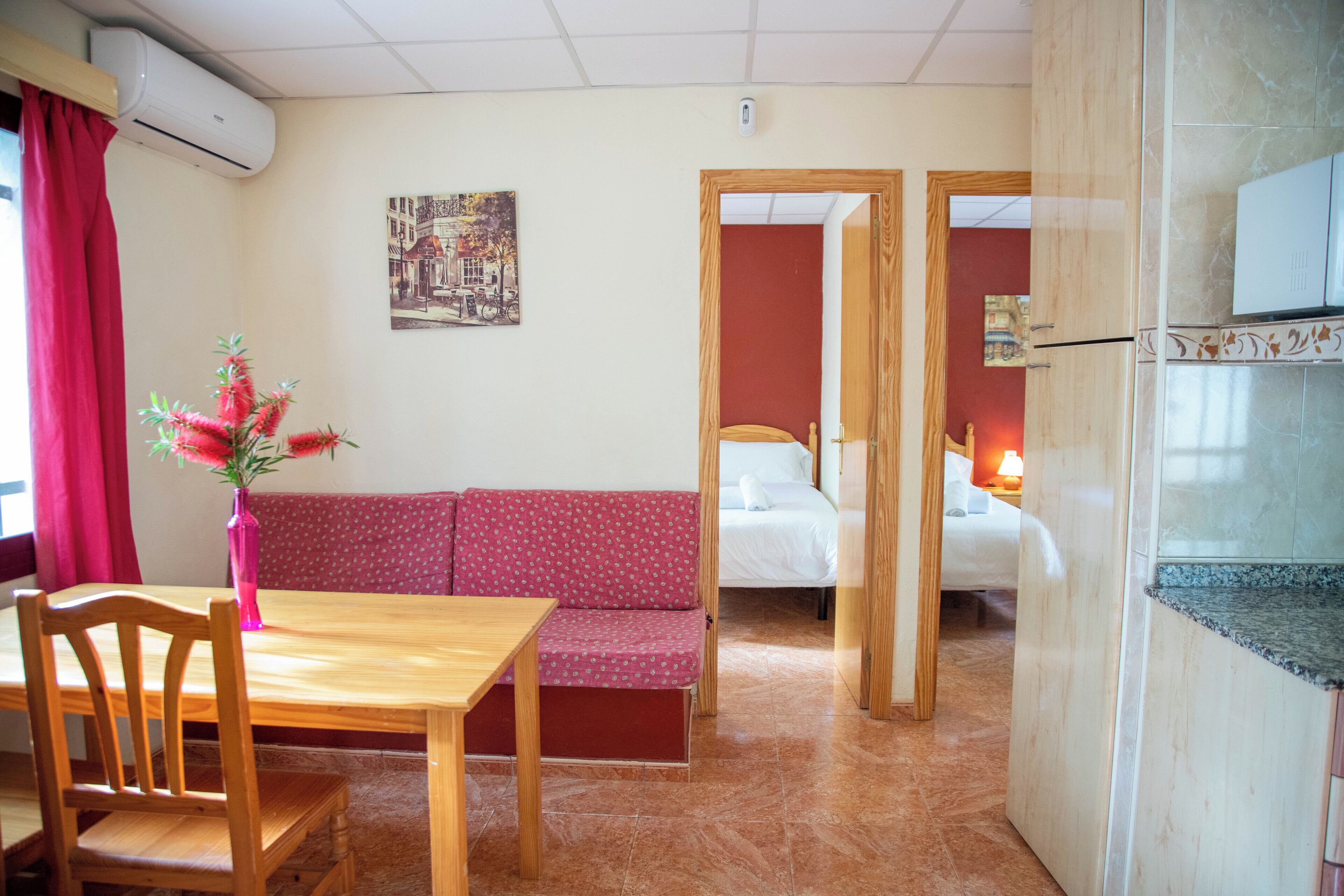 A hotel room with a wooden dining table, a pink sofa, a bed with white bedding, and a kitchen area with a microwave.