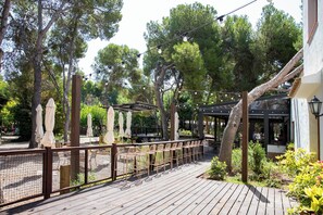 An outdoor patio with wooden tables and chairs, surrounded by trees and a pergola with string lights.
