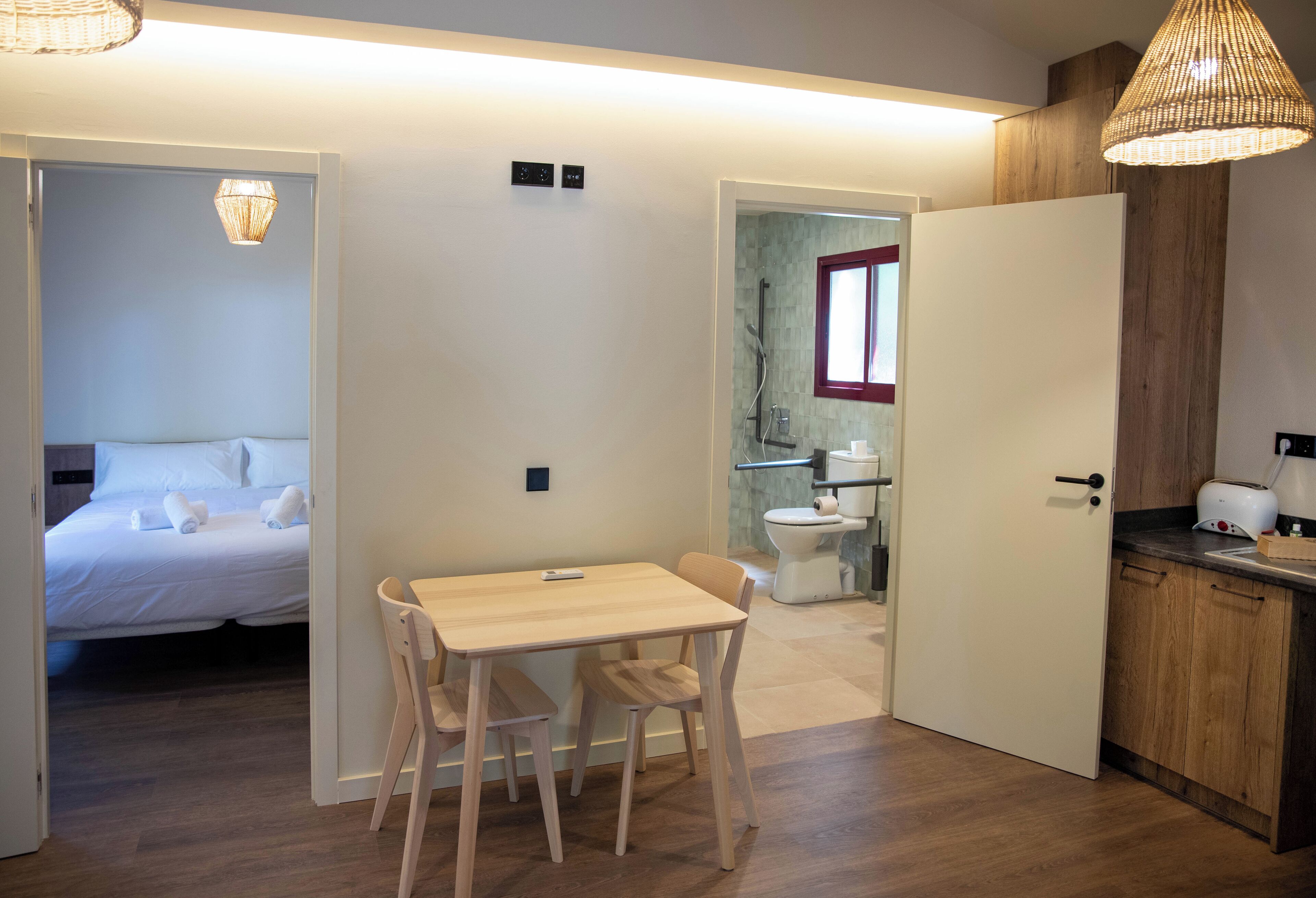 A compact hotel room with a bed, a small dining table with chairs, a kitchenette, and a bathroom with a toilet and sink.