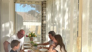 Un comedor con una mesa dispuesta para cuatro, una lámpara colgante y vistas al exterior a través de una ventana con cortinas traslúcidas.