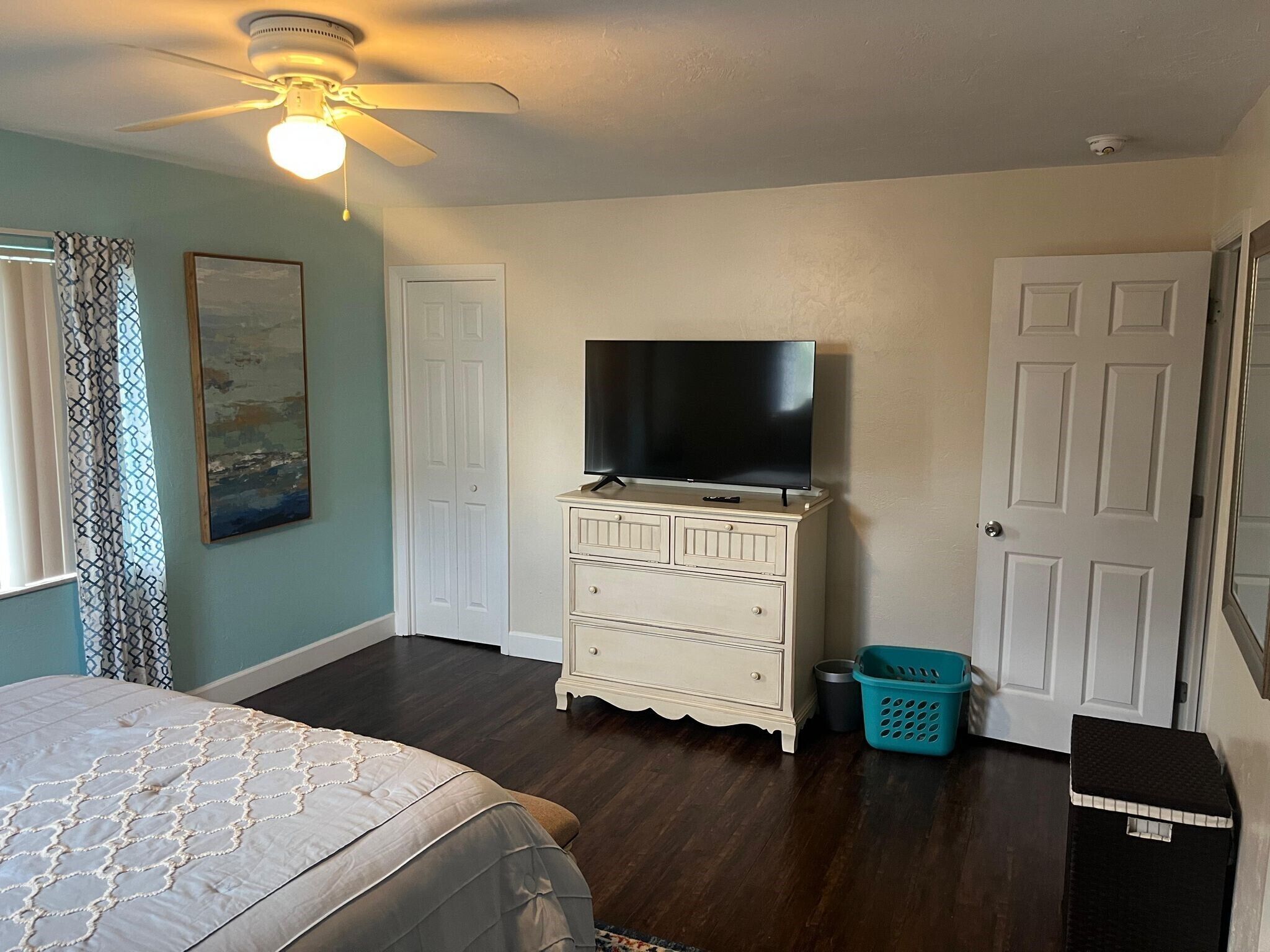 Ein Schlafzimmer mit Bett, Kommode, Fernseher, Deckenventilator und einer Tür.