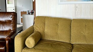 A living room with a brown leather armchair, a mustard-colored sofa, and a framed painting of a bird on the wall.