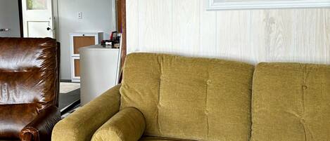 A living room with a brown leather armchair, a mustard-colored sofa, and a framed painting of a bird on the wall.