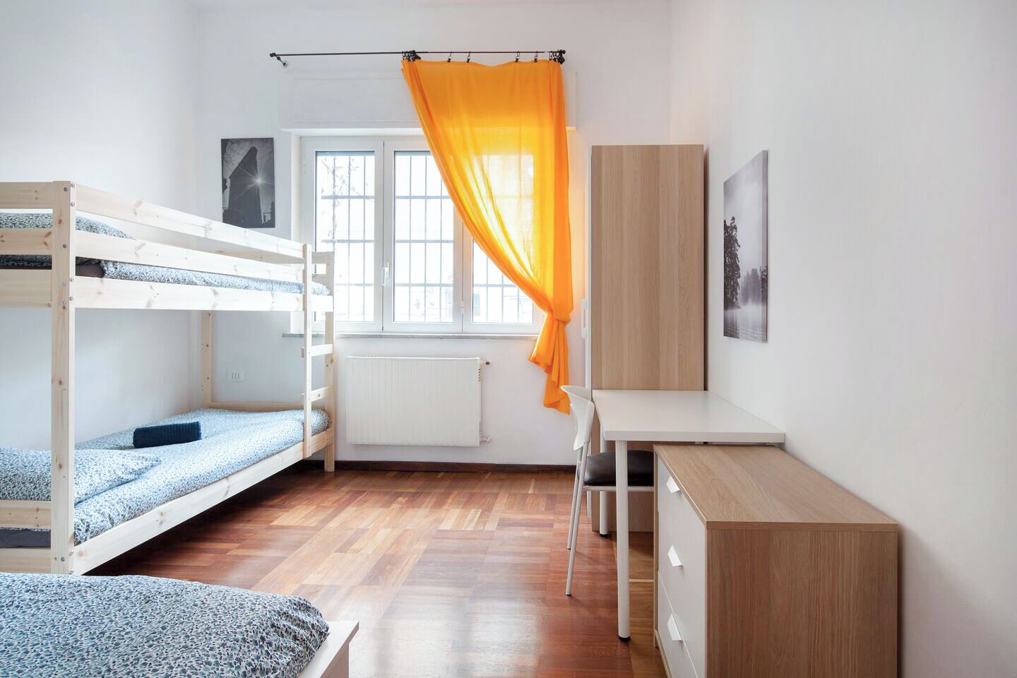 A dorm-style room with bunk beds, a desk, a chair, and a radiator.