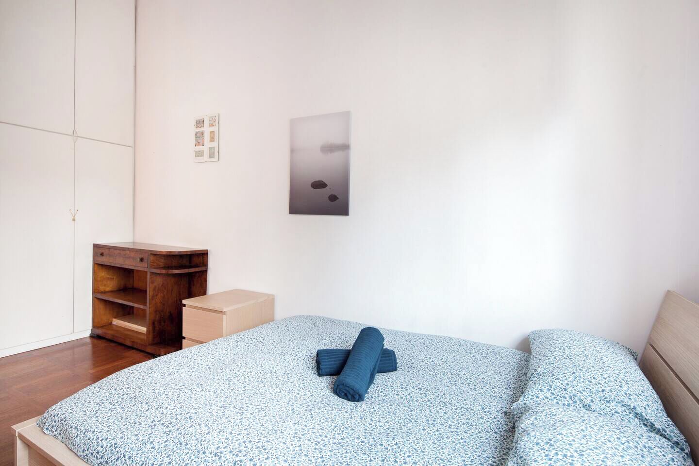 A bedroom with a bed, a wooden nightstand, a small wooden dresser, and a wall-mounted light fixture.