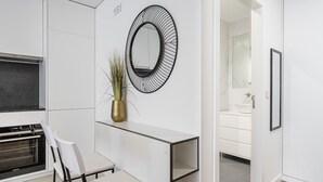 A modern kitchen with white cabinetry, a built-in oven, a dining area with a white table and chairs, a decorative mirror, and a potted plant.