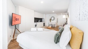 A modern hotel room with a large bed, a TV on a stand, a kitchen area, and a seating area with a table and chairs.
