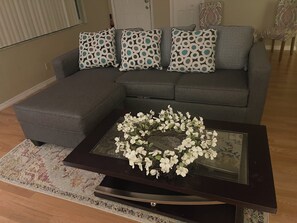 Un salon avec un canapé d’angle gris, une table basse en verre ornée de fleurs blanches et des coussins décoratifs à motifs.