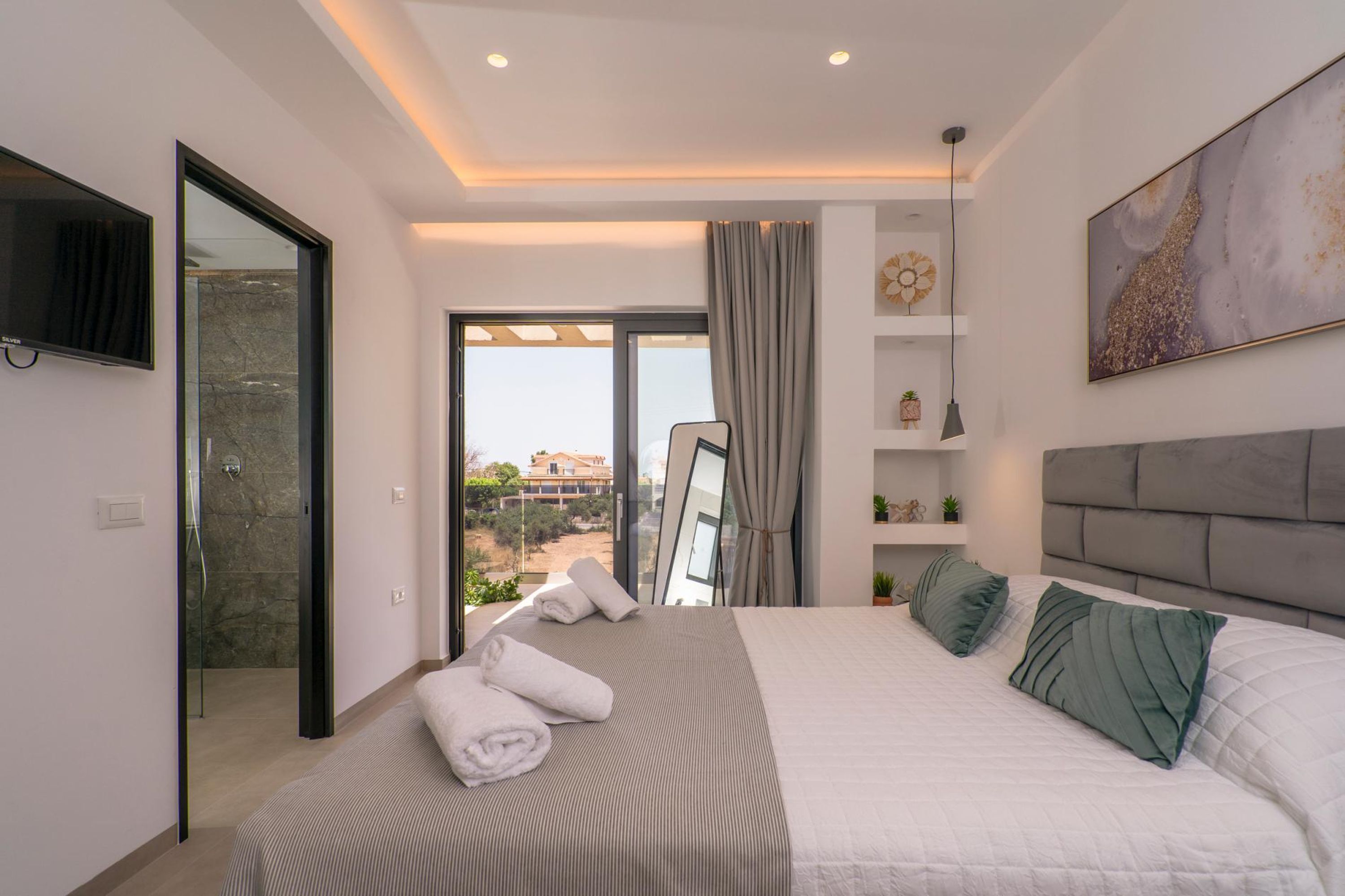 A modern bedroom with a large bed, a flat-screen TV, a sliding glass door to the balcony, and a view of the cityscape.