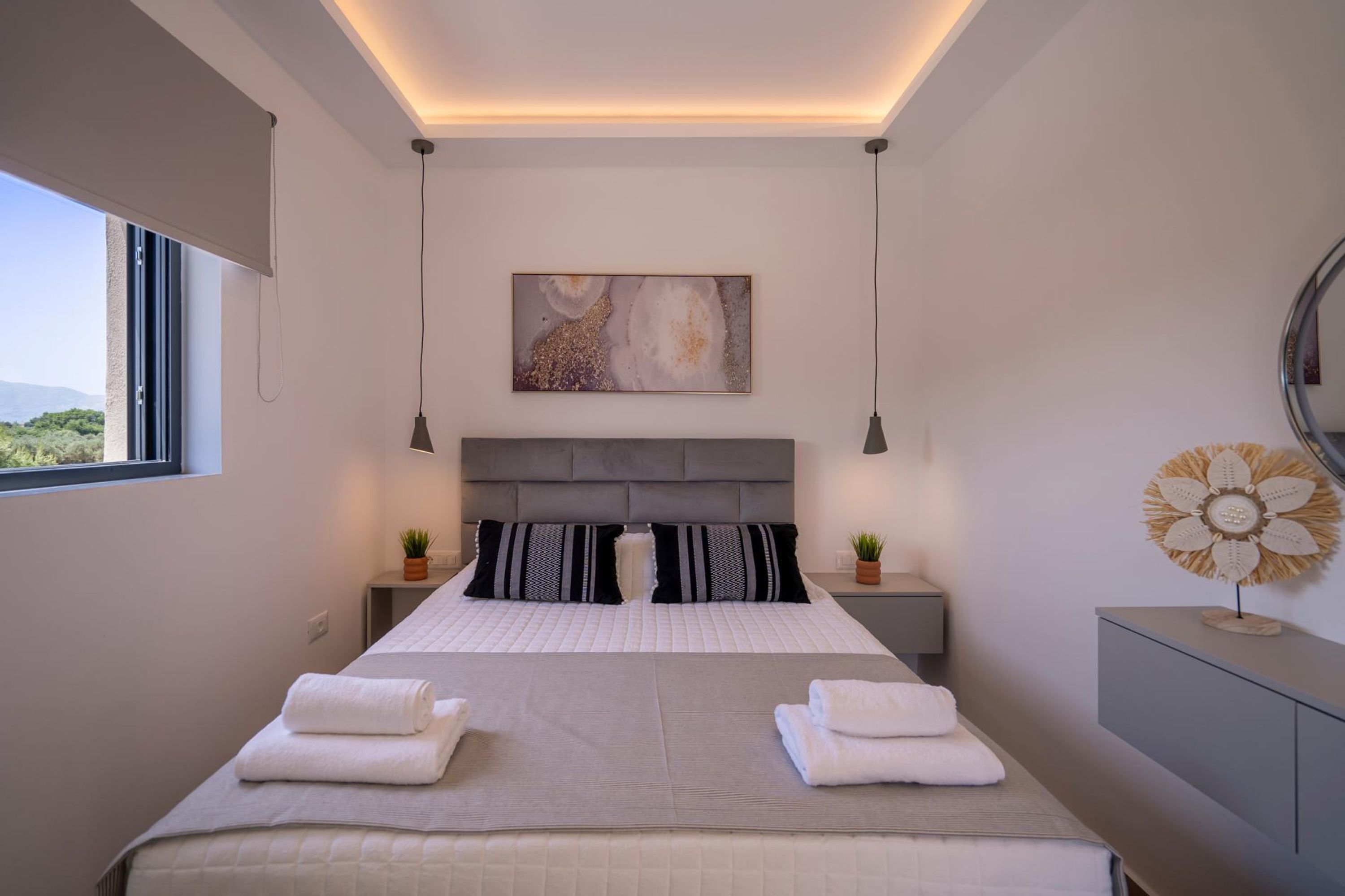 Ein modernes Schlafzimmer mit einem großen Bett, Nachttischen, einer Deckenleuchte, einem Wandbild und einem Fenster mit Blick.