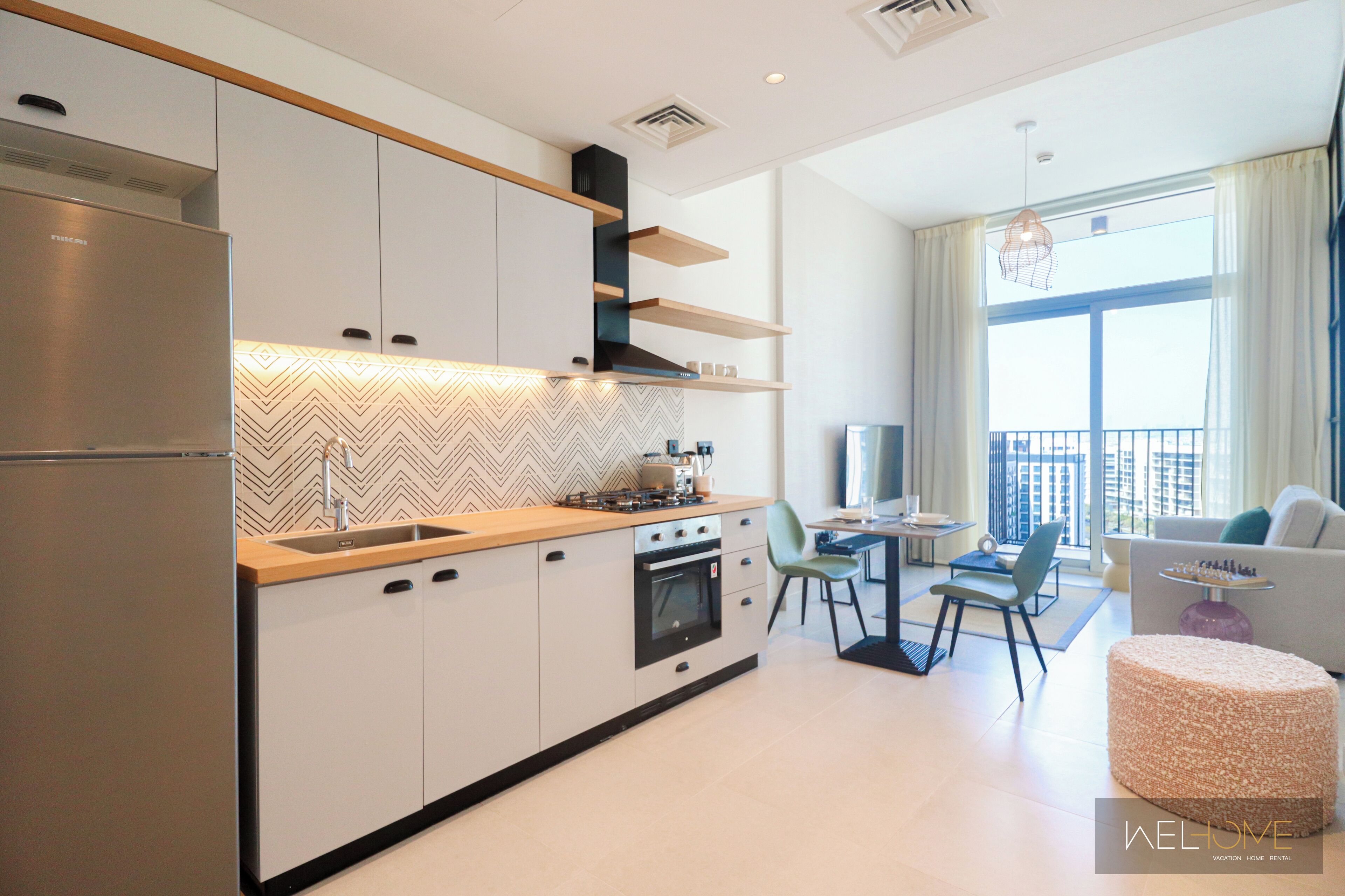 Een moderne keuken met een inbouwoven, een roestvrijstalen koelkast en een houten aanrecht. Er is een eethoek met een tafel en stoelen, en een bank in de woonkamer.