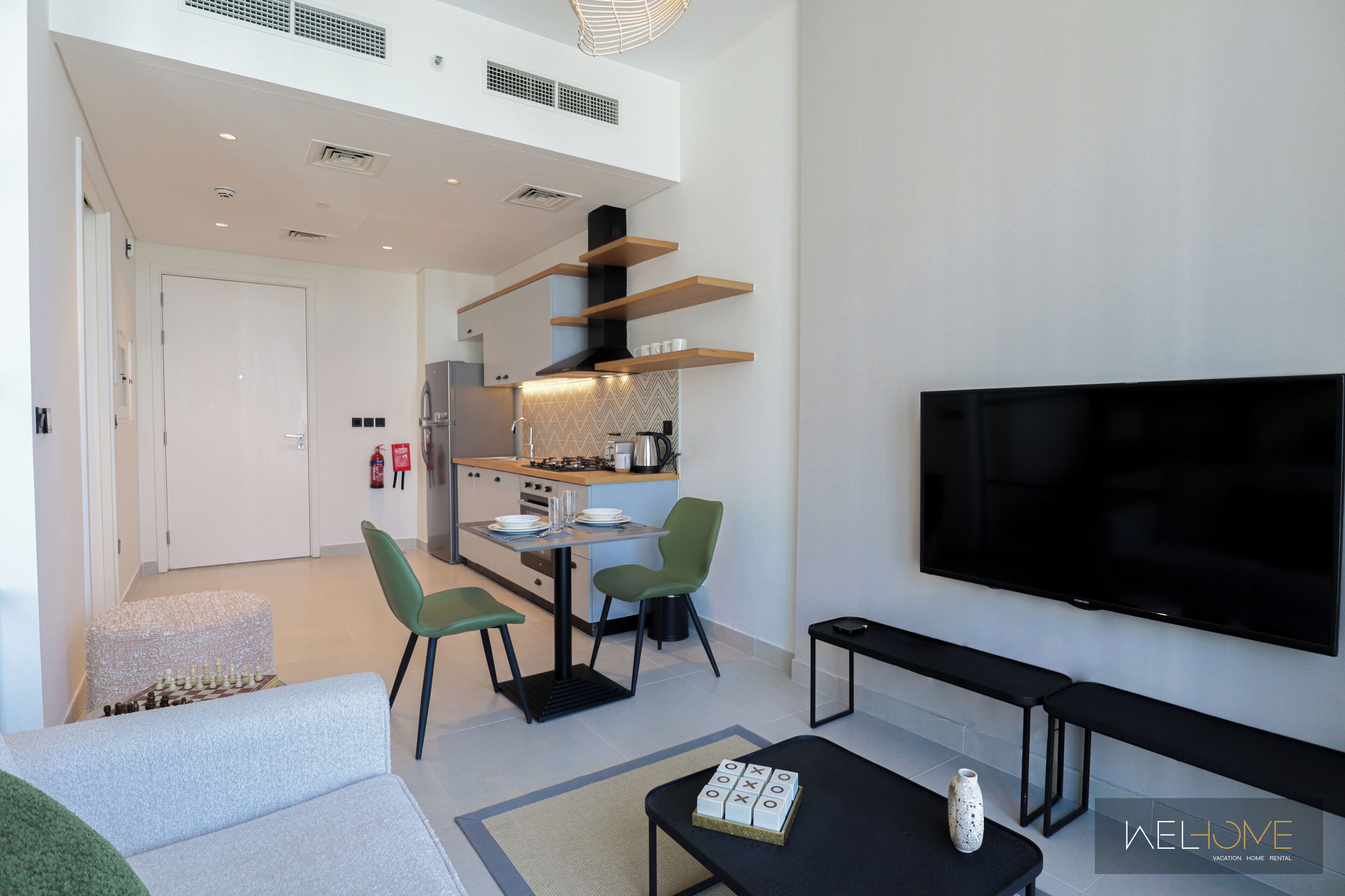 Una sala de estar moderna con televisor de pantalla plana, una mesa de centro y un sofá. Hay un comedor con una mesa y sillas, y una cocina al fondo.