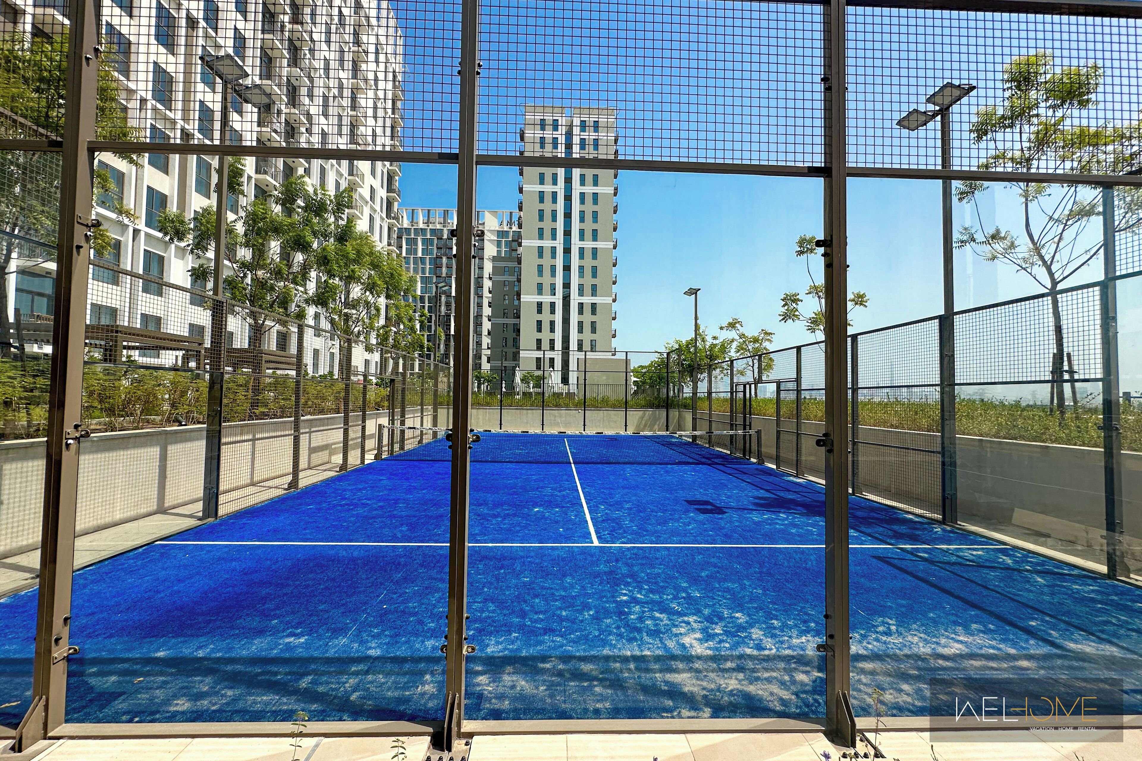 Ein Tennisplatz mit Netz, umgeben von einem Metallzaun und Gebäuden im Hintergrund.