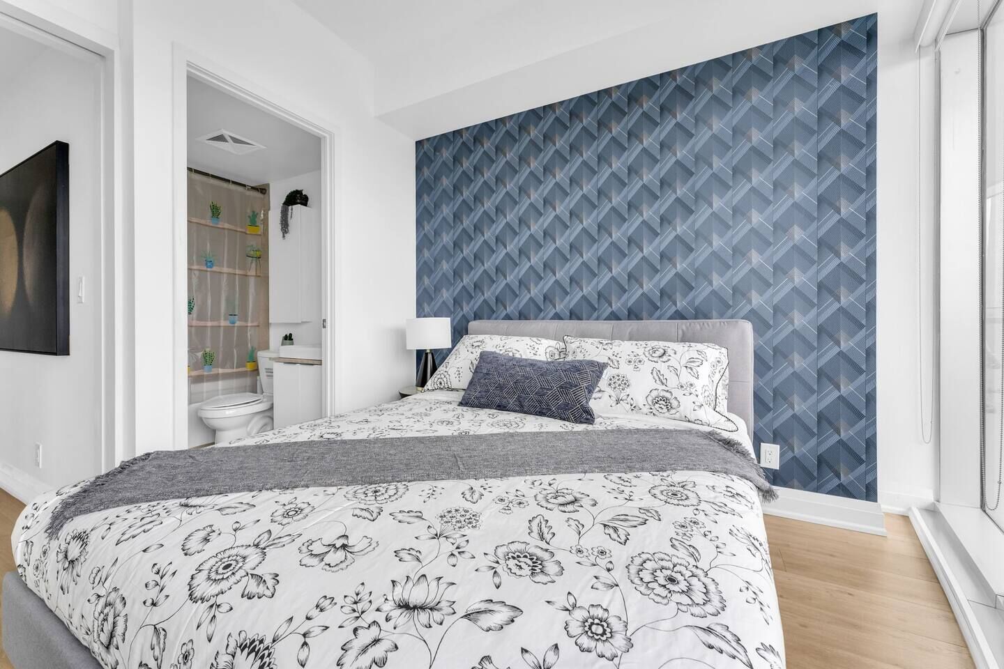 Een slaapkamer met een bed, nachtkastje, muur met een geometrisch patroon, raam en een badkamer zichtbaar door een open deur.