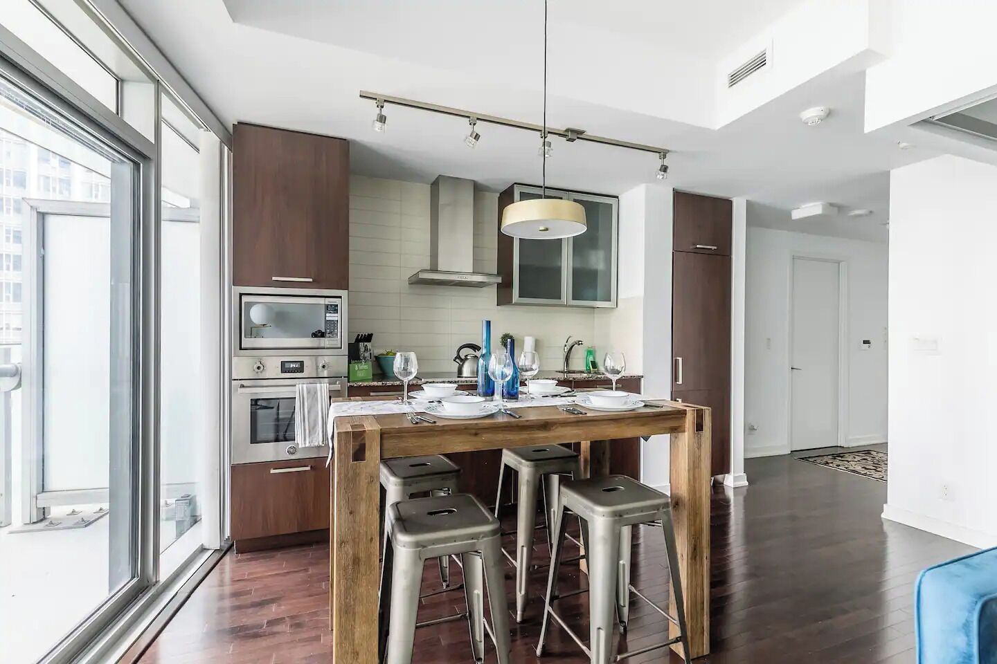 A modern kitchen with a wooden table, bar stools, and a hanging light fixture.