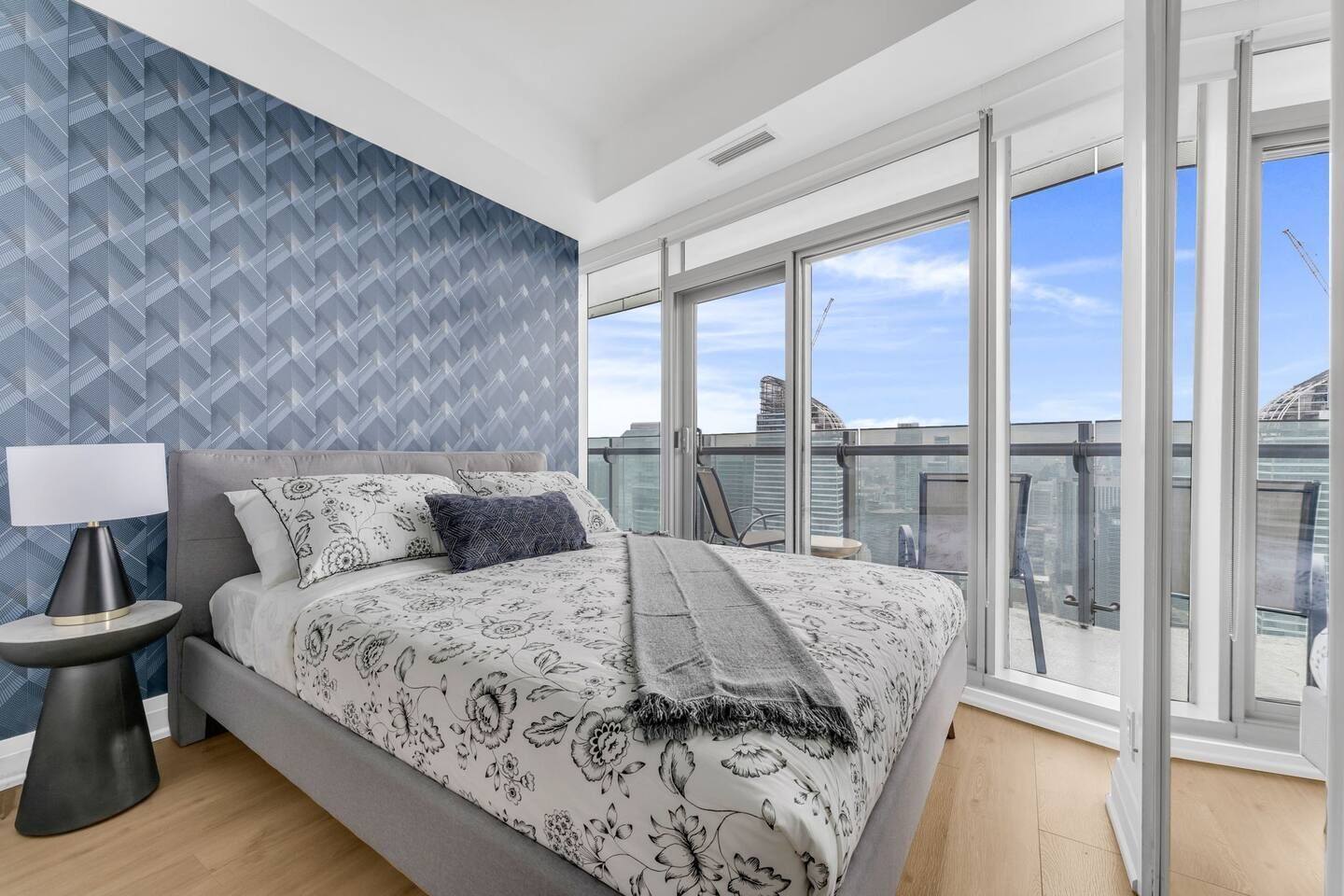 A modern bedroom with a large bed, a nightstand, a lamp, and a view of the city through large windows.