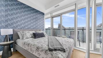 A modern bedroom with a large bed, a nightstand, a lamp, and a view of the city through large windows.
