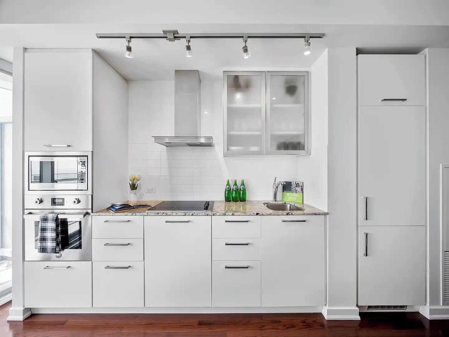 A modern kitchen with white cabinets, a built-in oven, a stove, and a microwave. There are three overhead lights and a window above the sink.