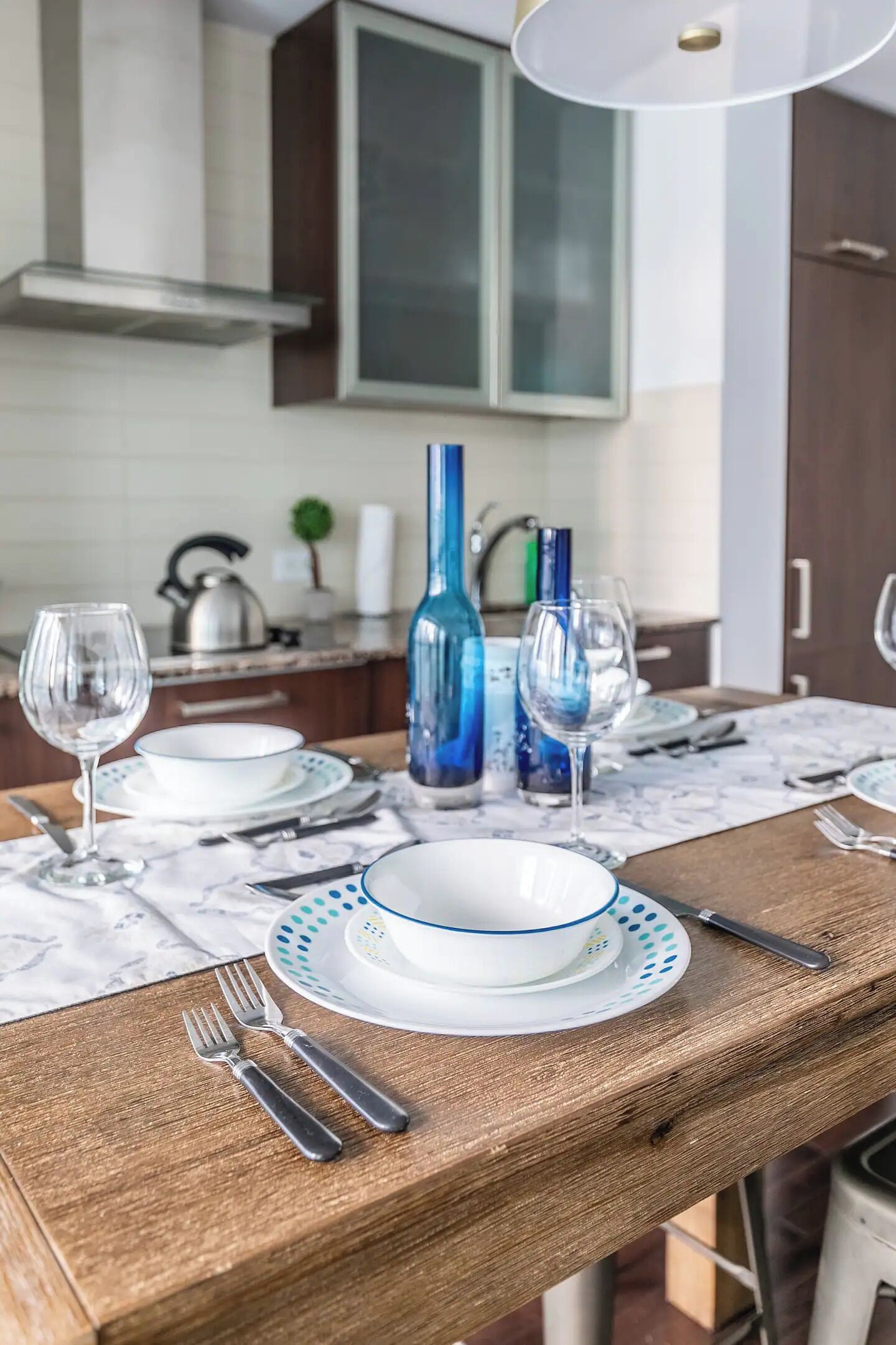 Een eettafel gedekt met borden, glazen en bestek, met een blauwe vaas en een waterkoker op de achtergrond.