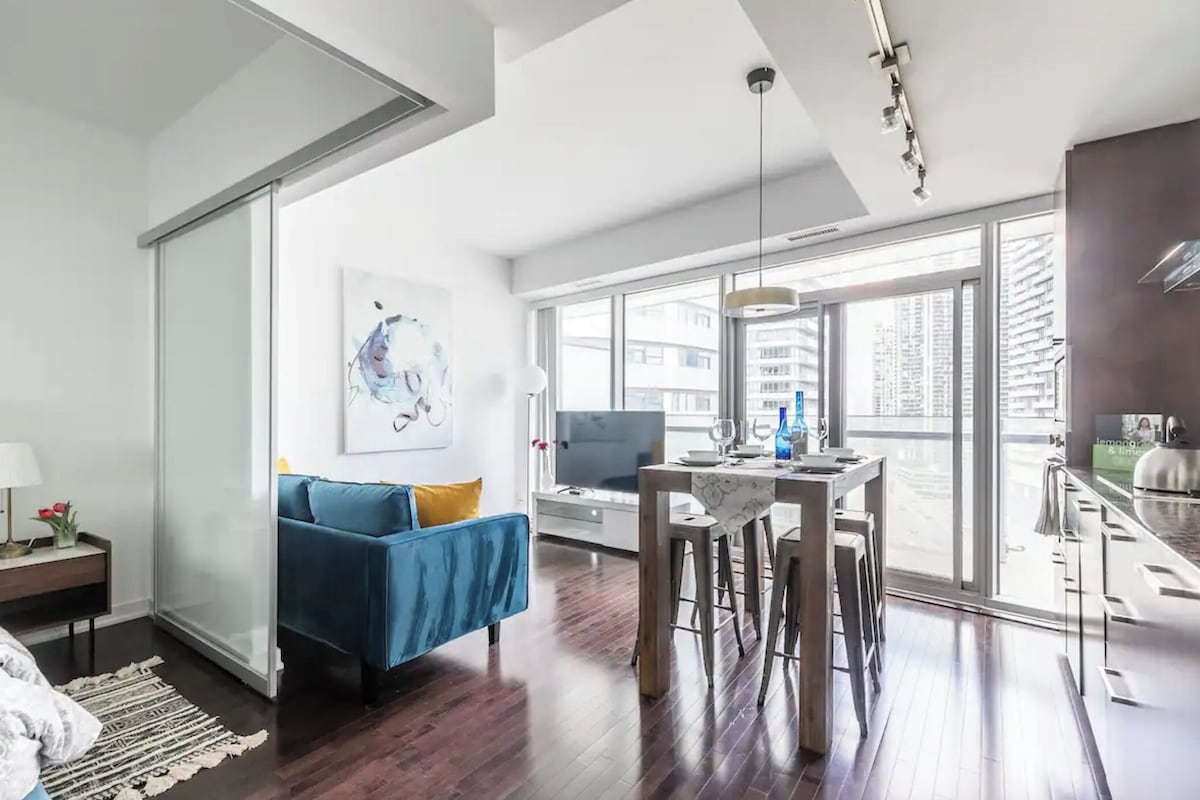 Una sala de estar moderna con un sofá azul, un comedor con una mesa y sillas, y ventanas amplias que ofrecen vistas de la ciudad.