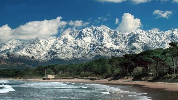 Paisagem costeira com montanhas nevadas ao fundo, uma praia de areia e uma pequena construção perto da água.