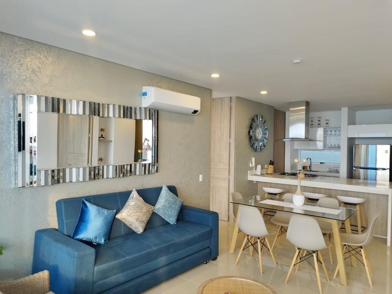 Un salon moderne avec un canapé bleu, un coin repas doté d’une table en verre et une cuisine équipée d’appareils électroménagers en acier inoxydable.