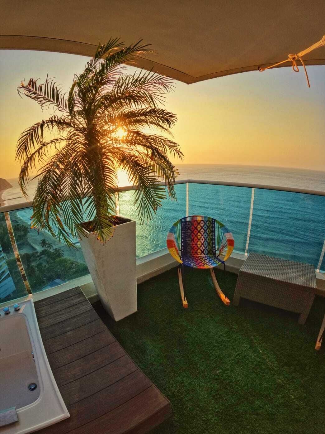 A balcony with a palm tree, a colorful chair, a small table, and a view of the ocean.
