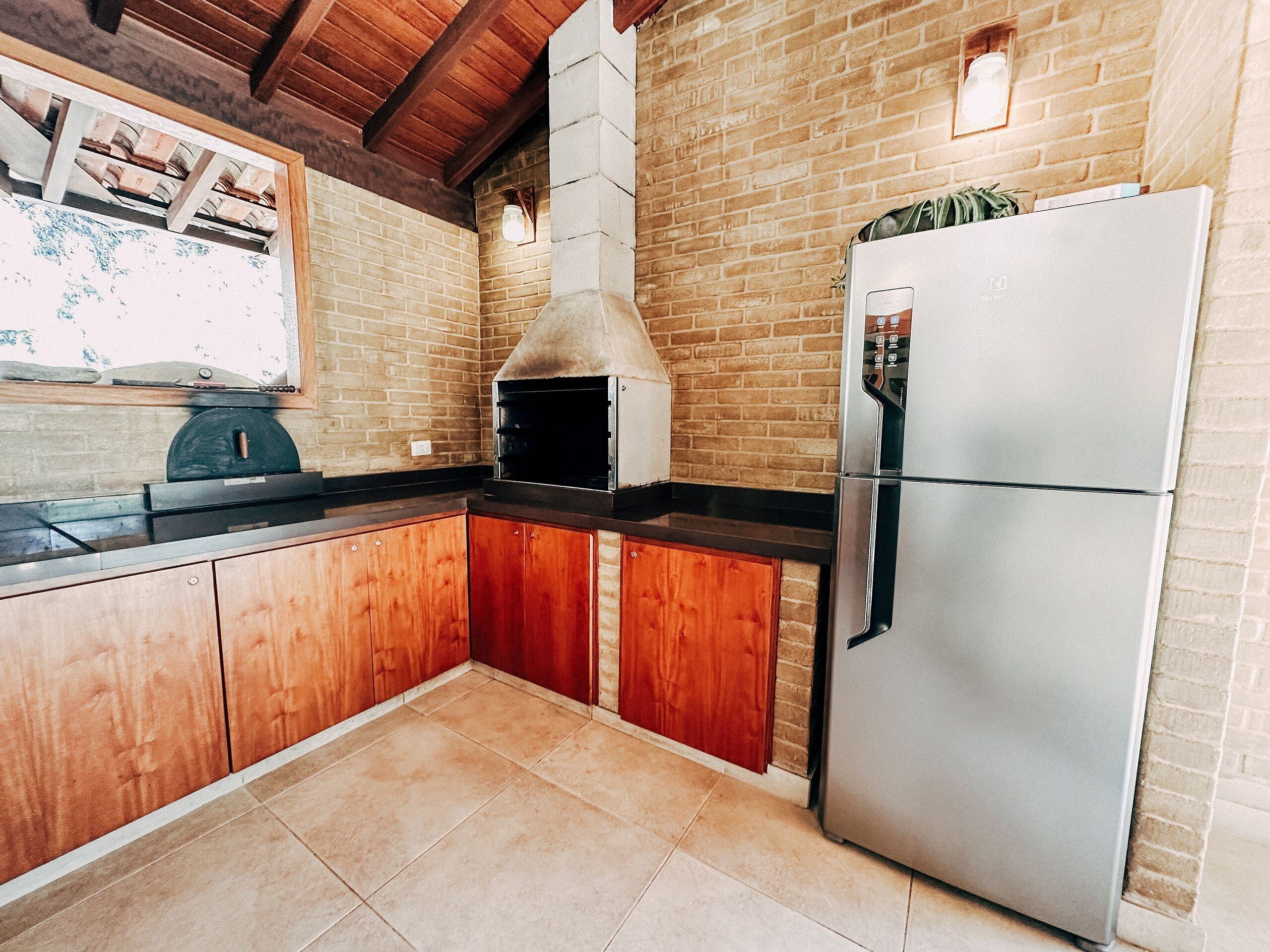 A kitchen with wooden cabinets, a brick wall, a stainless steel refrigerator, and a pizza oven.