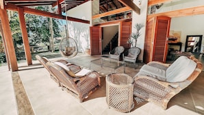 A covered outdoor seating area with wicker furniture and a glass table, surrounded by a wooden pergola and a view of a staircase and greenery. - Casa Rio Habitat (Petrópolis)