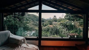 A room with a large window offering a view of a lush, green landscape with hills and a river. - Casa Rio Habitat (Petrópolis)