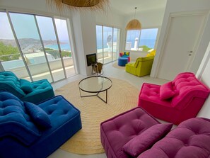 A spacious living room with large windows offering a view of the sea and hills. There are colorful sofas and a round coffee table.