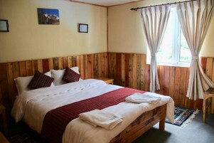 Quarto com cabeceira de madeira, cama com roupa de cama branca e detalhes vermelhos, janela com cortinas e uma pequena mesa de cabeceira.